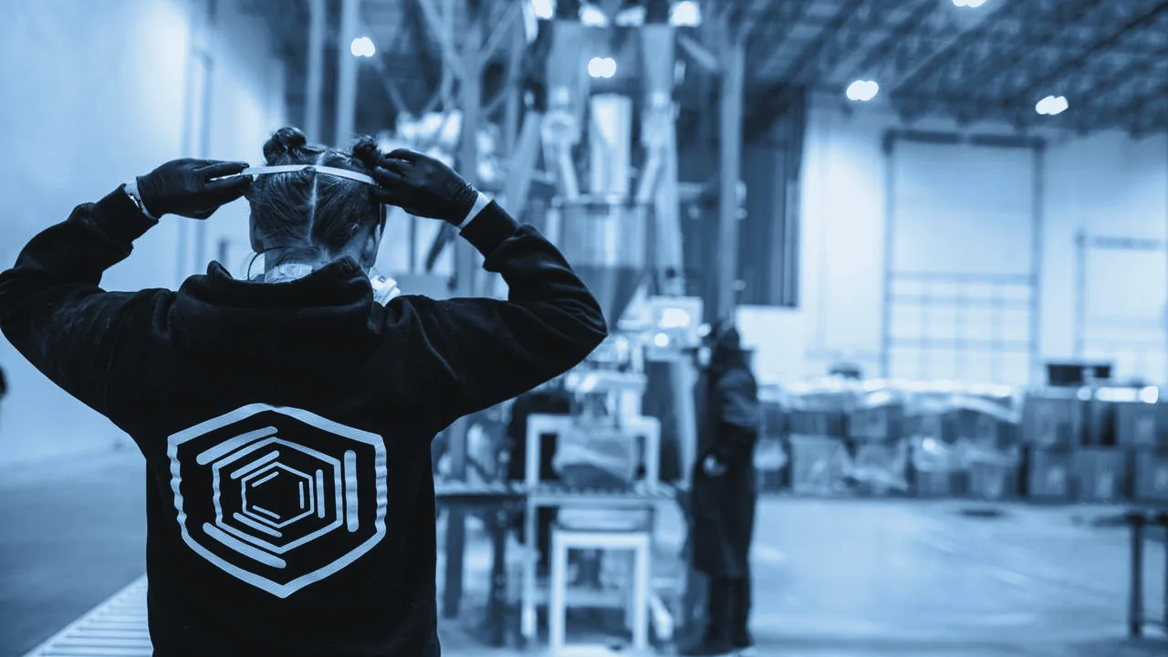 Worker seen from behind tying on a face mask on a production floor, with equipment behind