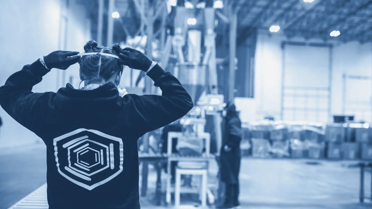 Worker seen from behind tying on a face mask on a production floor, with equipment behind