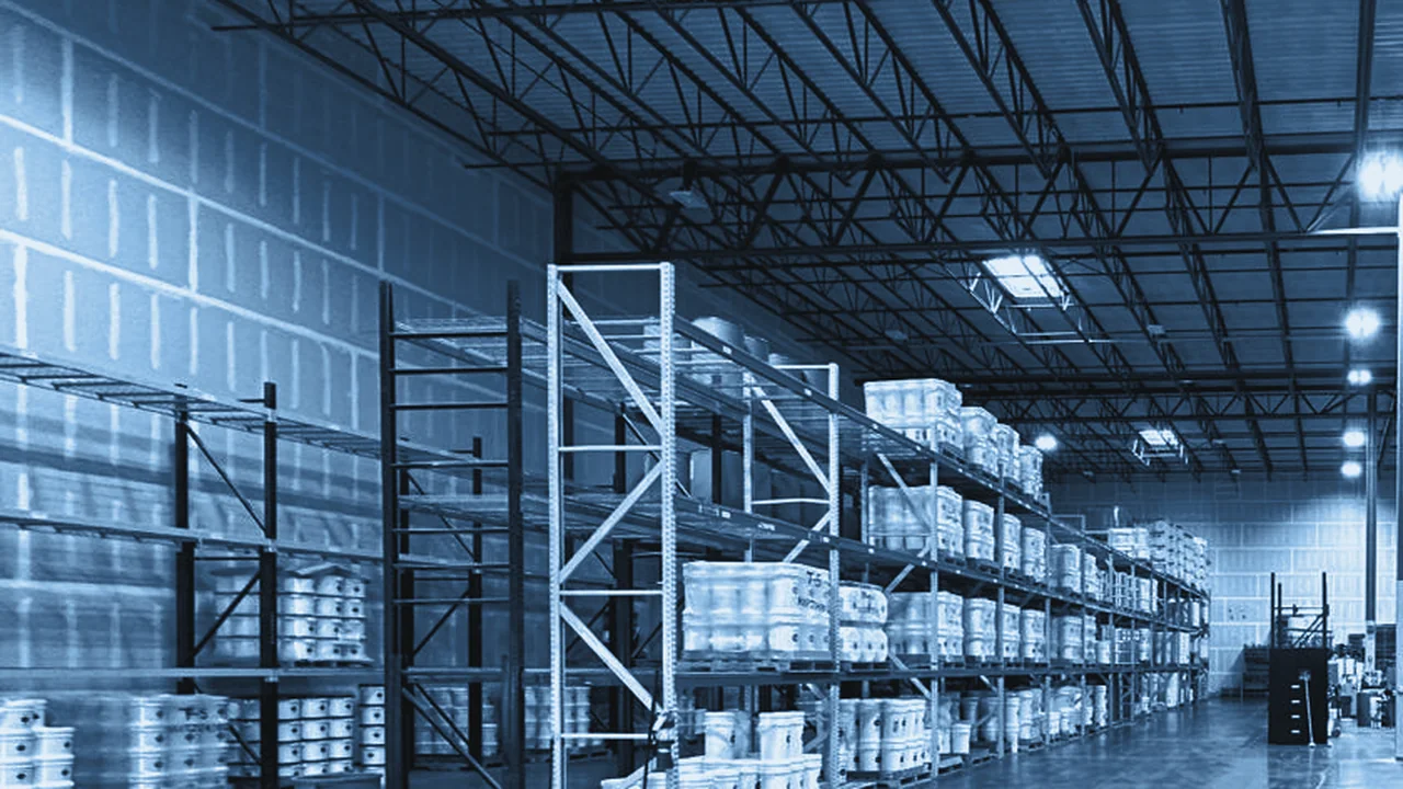 Open warehouse floor with tall pallet racks of white pails under a steel truss ceiling
