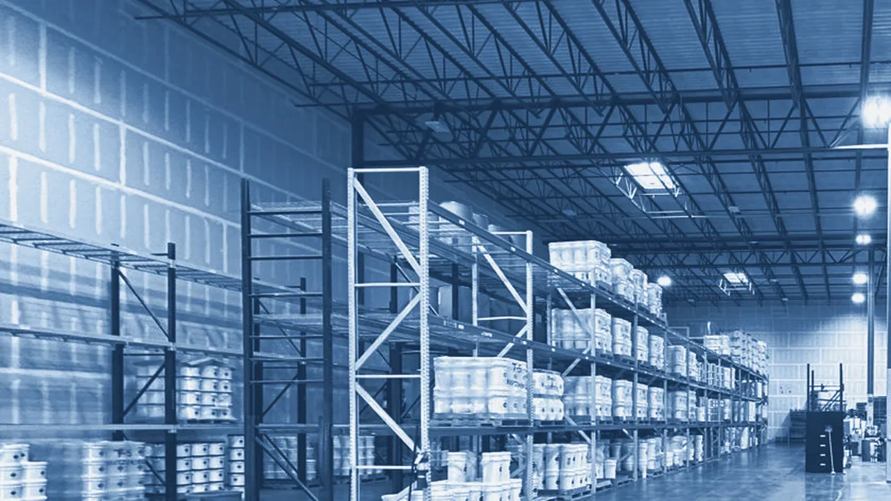 Open warehouse floor with tall pallet racks of white pails under a steel truss ceiling